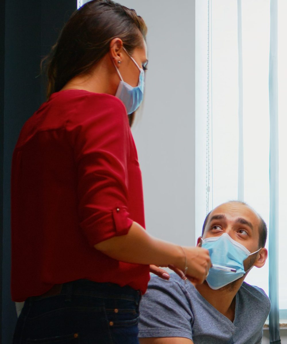 Colleagues with masks discussing graphics sitting in new normal office room. Coworkers talking working pointing at desktop respecting social distancing against covid virus using plexiglass.